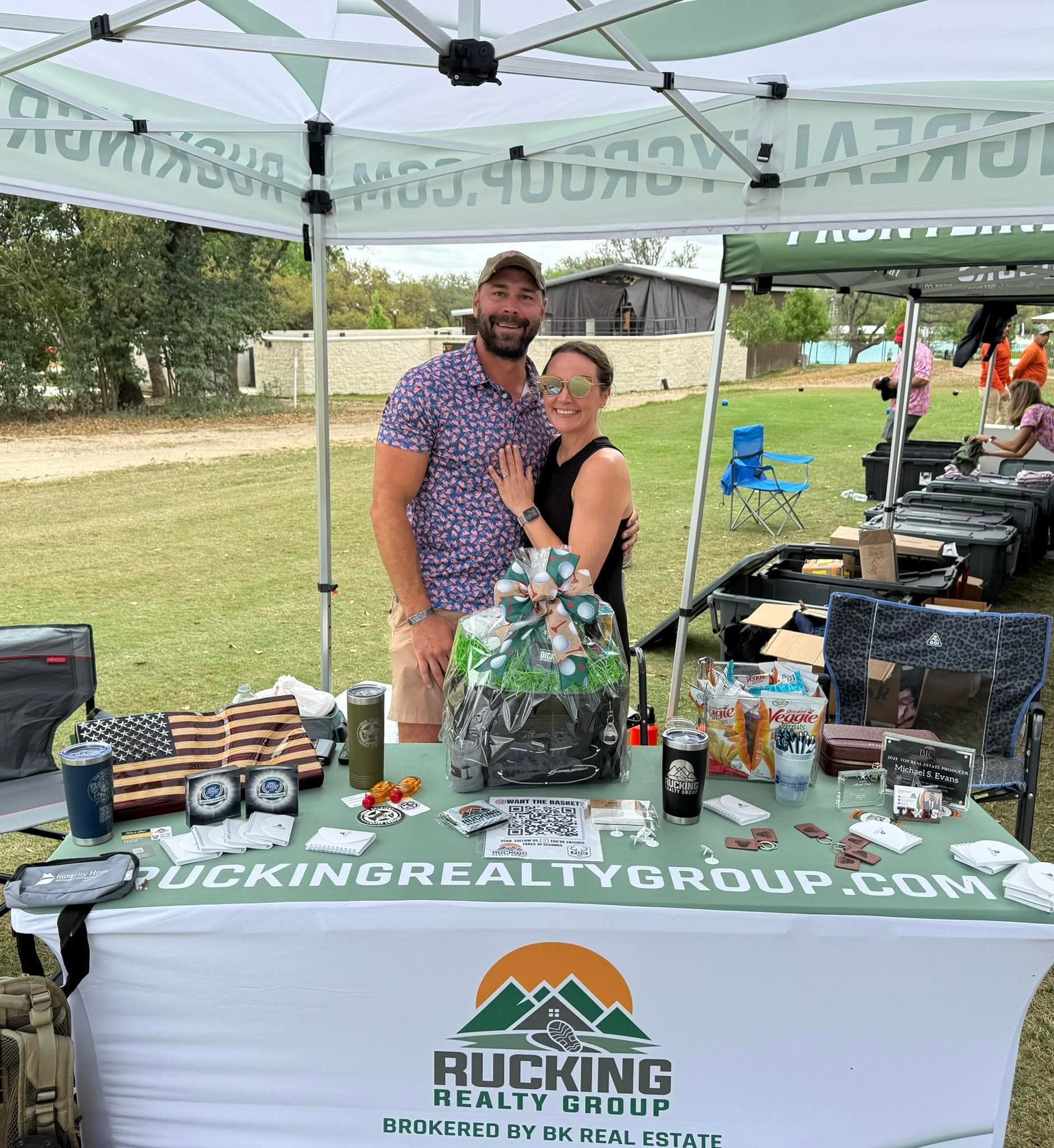 Mike and Natalie Evans under the Rucking Realty Group tent at the Frontline Hero Outdoors golf tournament, beside a table with a Rucking Realty Group cover