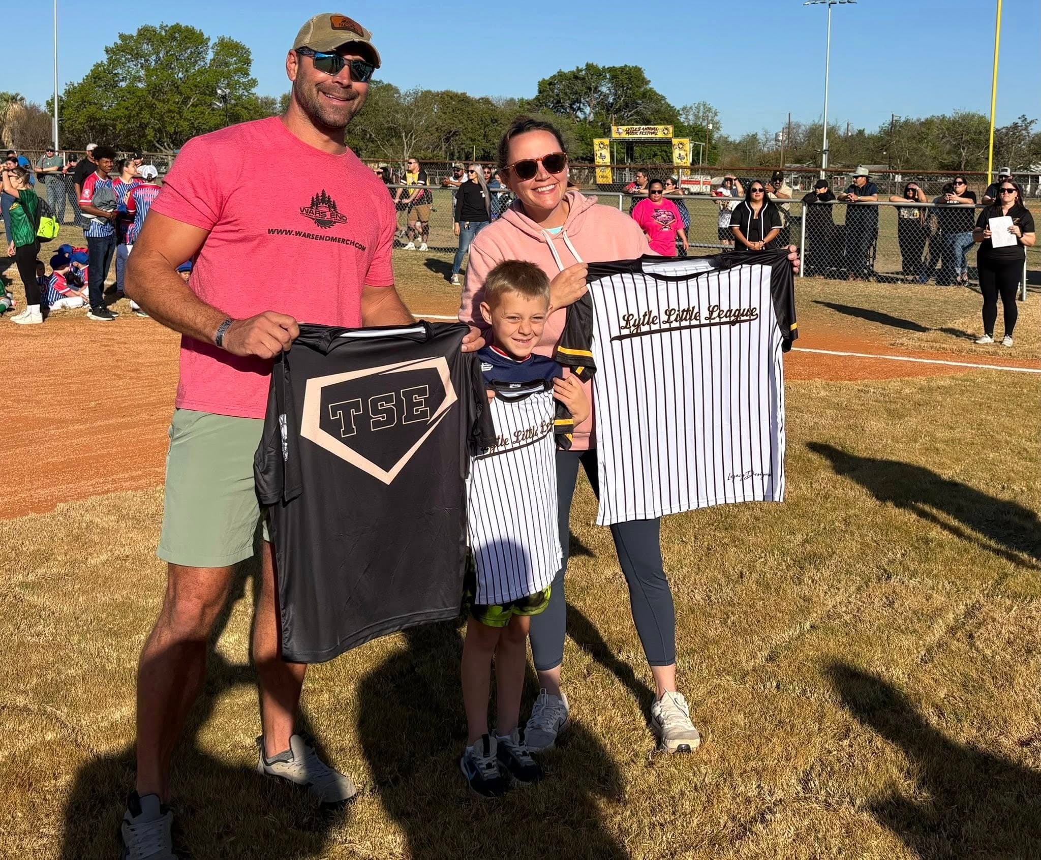 Mike and Natalie Evans and their son on a ballfield, holding up Lytle Little League jerseys