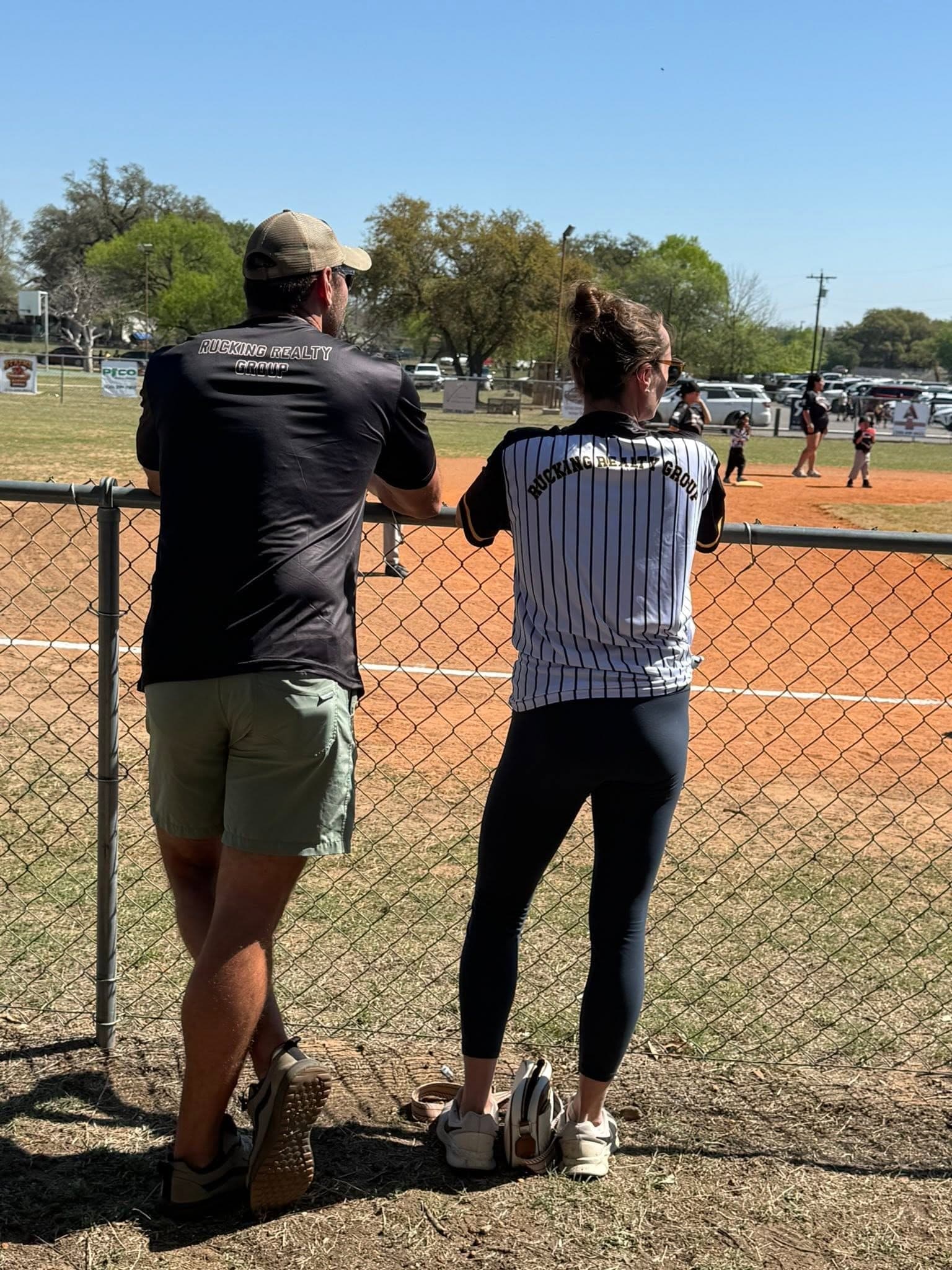Mike and Natalie Evans in Rucking Realty Group shirts, leaning on the outfield fence watching the game